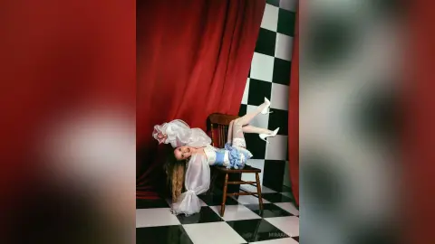 Fashionable woman in a white blouse and blue skirt reclining playfully on a chair against red curtains.