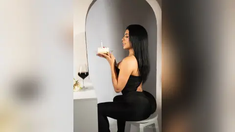 Elegant woman in a black jumpsuit holding a small cake in a minimalist white room for a celebratory mood.