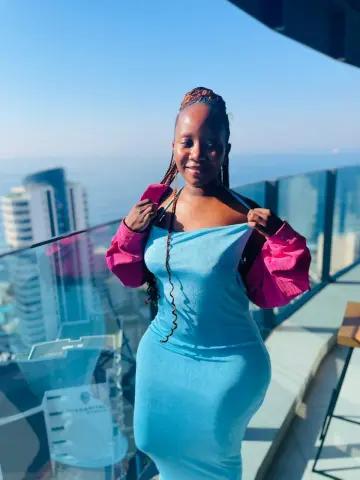 Smiling woman in a blue midi dress posing on a high-rise balcony overlooking a city skyline under a clear sky.