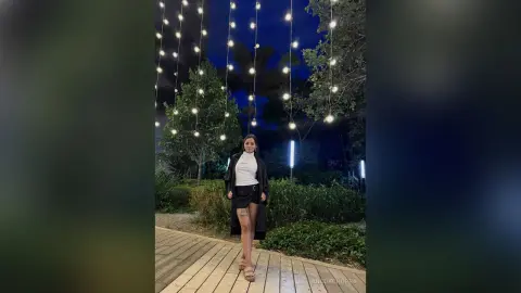 Stylish woman in a white turtleneck and black skirt standing under fairy lights in a lush evening garden.