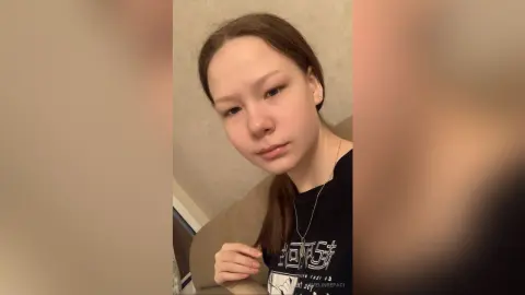 Young woman with long brown hair wearing a black graphic t-shirt in a cozy indoor setting.
