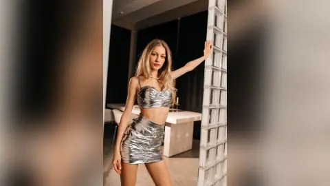 Glamorous blonde woman in a metallic silver crop top and mini skirt posing in a modern minimalist room.