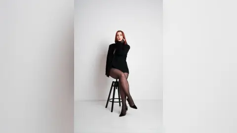 AryaSin poses sitting on a stool wearing a black long-sleeve top and black tights or pantyhose against a white background.