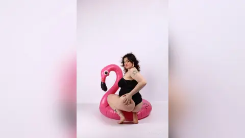 KiraKovall poses naked behind a pink flamingo float, wearing a black bra and showing her bare buttocks and legs against a white background.