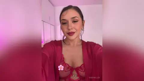 Smiling woman in a pink lace bodysuit and matching robe posing in a bright, modern room.