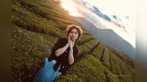 Young woman in a black top and blue skirt sitting in a lush green tea plantation at sunset.