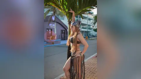 AndreaKalo poses outdoors in a black bikini top and a long, fringed black skirt, standing next to a palm tree on a street.