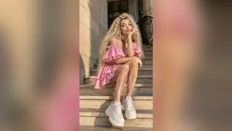 Blonde woman in a pink ruffled mini dress and white platform sneakers sitting on stone stairs in warm sunlight.