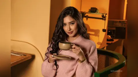 Young woman in a pink knit sweater enjoying coffee in a warm, cozy indoor setting.