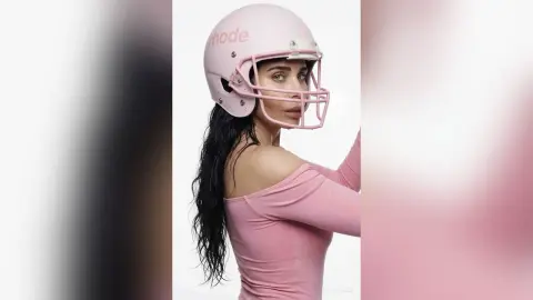 Fashionable woman in a pink off-the-shoulder top and matching pink football helmet against a white background.