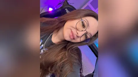 Smiling young woman with long brown hair and glasses in a purple lit gaming room setting.