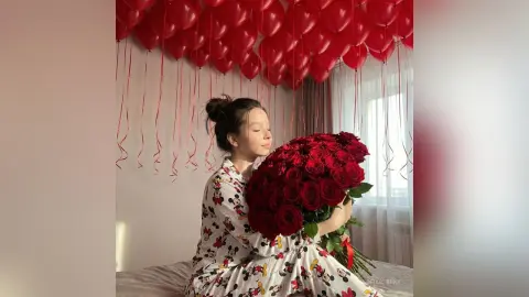 Happy woman in floral dress hugging a large bouquet of red roses under floating red balloons.