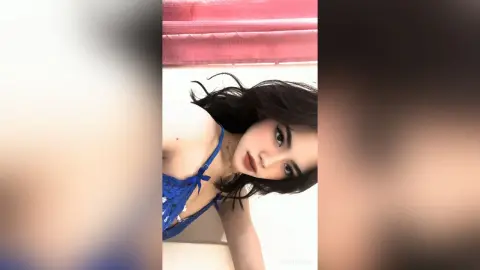 NancyLoppez poses in a blue lace bra with her dark hair falling over her shoulders against a light background.
