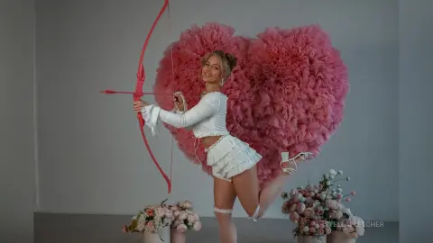 StellaFletcher poses in a white lace crop top and skirt, holding a red bow and arrow against a large pink heart-shaped feather backdrop.