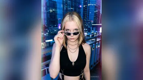 JaimieBez wears silver sunglasses and a black halter top while standing on a balcony overlooking a city at night.