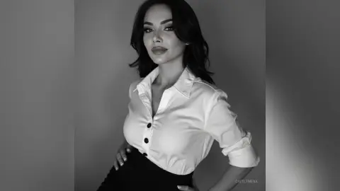 Elegant woman in a white button-down shirt posing for a sophisticated black and white studio portrait.