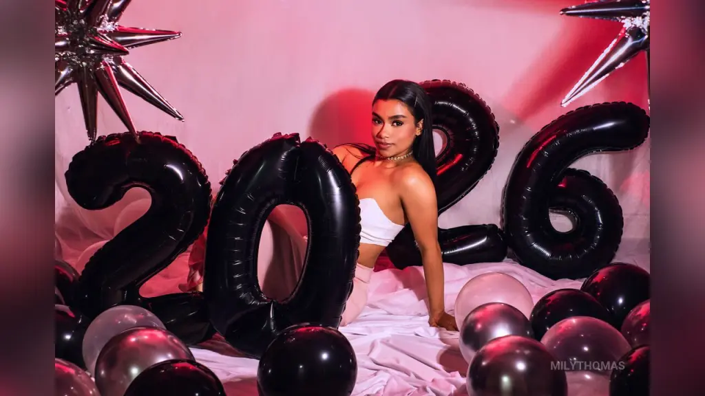 MilyThomas β Stylish woman in white outfit sitting by large black 2026 balloons for a celebratory New Year's Eve mood.