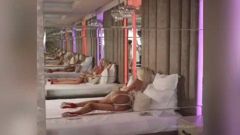 Blonde woman in white lingerie relaxing on a bed in a luxurious mirrored bedroom with warm lighting.