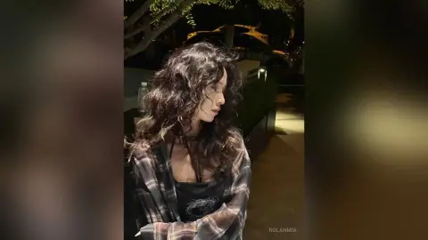 Woman with wavy hair wearing a plaid shirt in a moody nighttime outdoor setting.