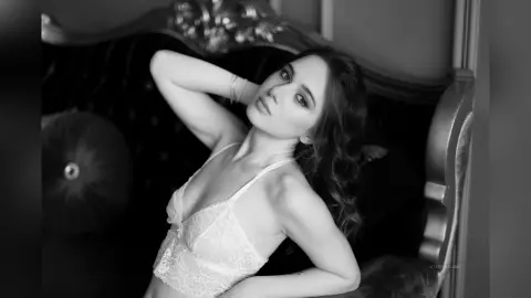 StacyHarland poses in white lace lingerie while leaning back against a black tufted sofa.