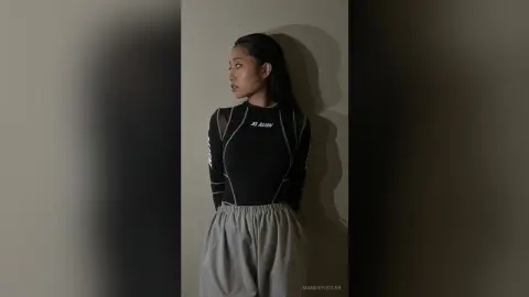 Young woman in a black long-sleeve crop top and grey joggers posing against a neutral wall in moody lighting.
