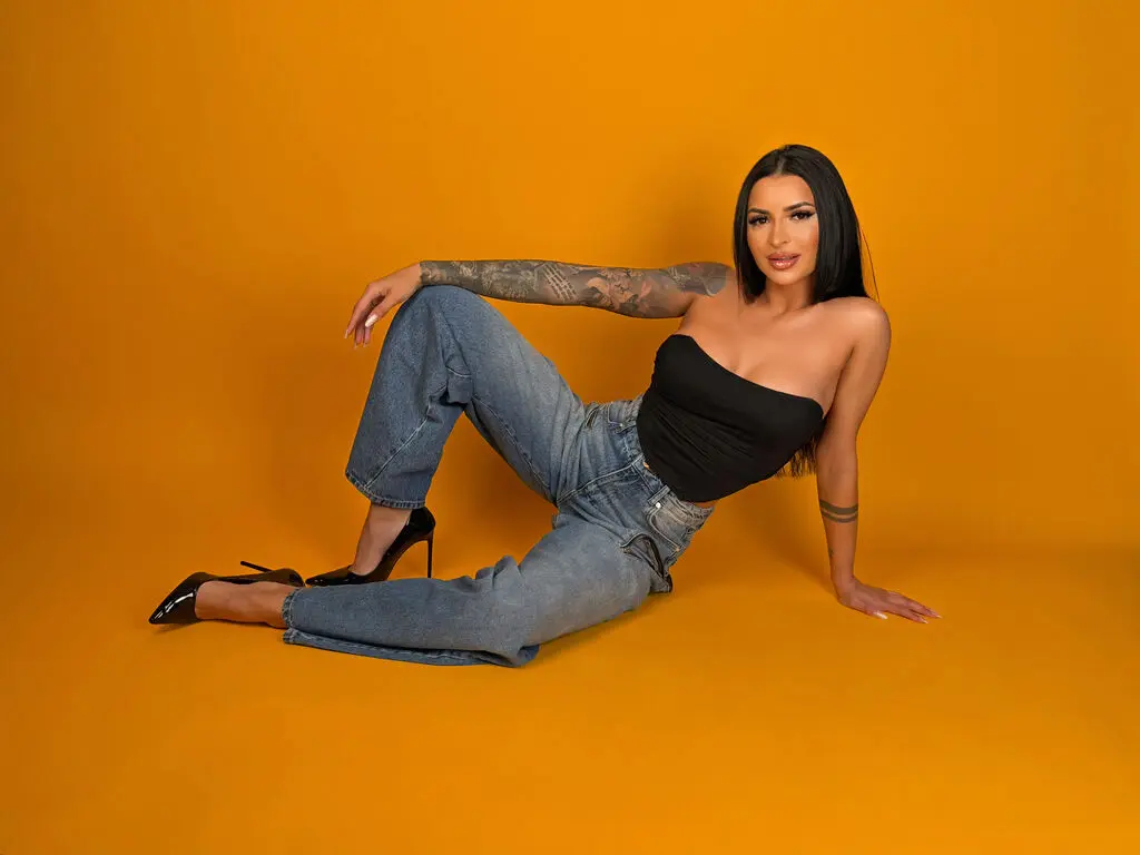 Tattooed woman in black tube top and blue jeans sitting against a vibrant yellow background.