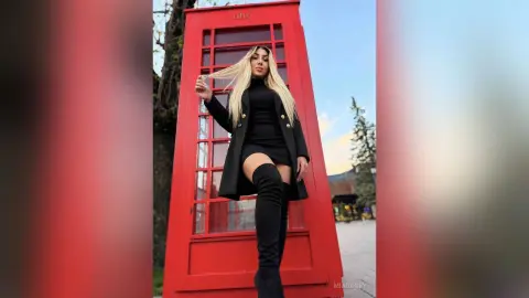 Stylish blonde woman in a black mini dress and over-the-knee boots posing by a red London telephone booth.