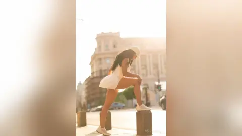 NicoleBella poses outdoors in a white bodysuit and white cap, leaning forward with one leg bent while looking down.