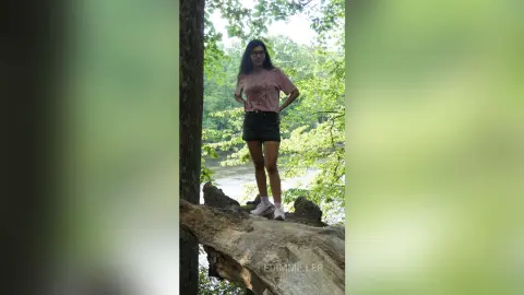 Young woman in a pink t-shirt and black shorts standing on a fallen log by a peaceful forest river.