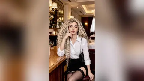 Elegant woman with long blonde curly hair in a white blouse and black skirt sitting at a luxury restaurant bar.