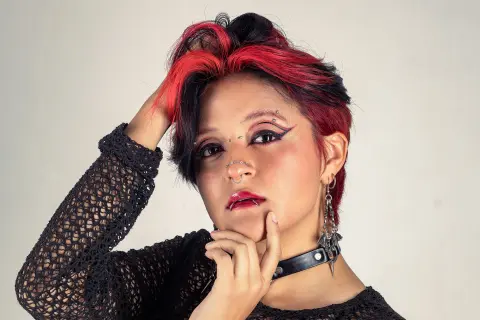 YuuMiko poses with her hand to her hair, showing her red and black hair, winged eyeliner, nose ring, and black mesh top.