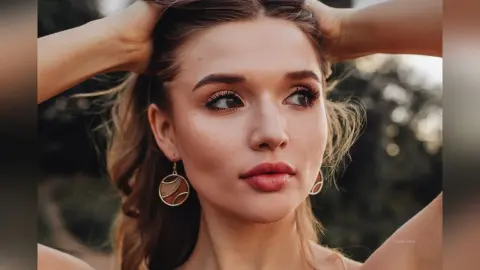 Portrait of a young woman with brown hair and gold earrings looking thoughtfully in a soft outdoor setting.