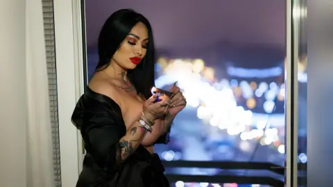 DeeaAnderson poses in a black satin robe, looking down while holding a lighter near her cleavage against a blurred night city background.