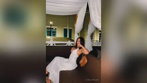 Young woman in a white summer dress sitting on a patio sofa at night under a canopy.