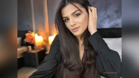 Young woman with long dark hair wearing a black blouse in a warm, dimly lit indoor setting.