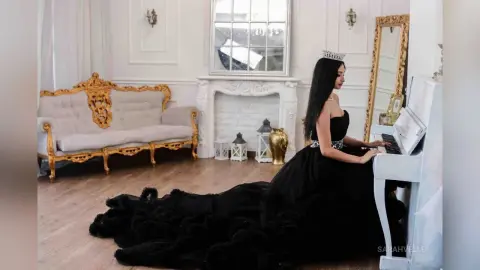 SarahVelle sits at a white piano wearing a black strapless ballgown and a silver tiara.
