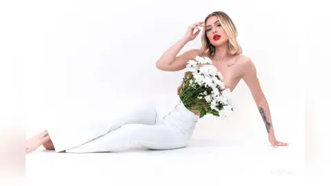DaisyLin reclines on a white floor, holding a large bunch of white flowers against her bare chest and torso while wearing white trousers.