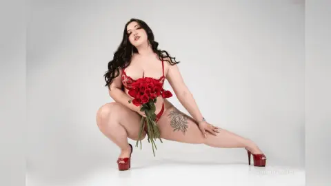 Curvy woman in red lace lingerie holding a bouquet of red roses in a bright studio setting.