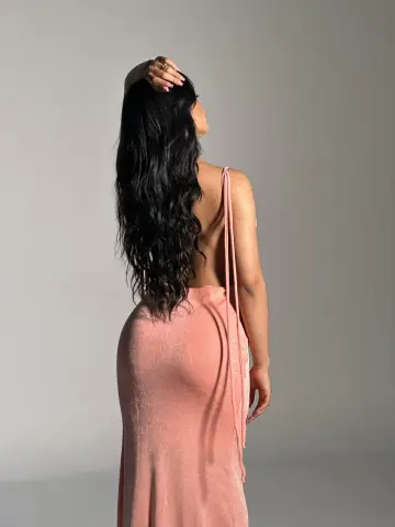 Woman with long wavy black hair wearing a backless pink maxi dress in a minimalist studio setting.