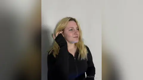 StellaVanilla, a blonde woman, wearing a black long-sleeved top, tucking her hair behind her ear against a plain white wall.