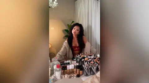 Young woman in a cozy cream cardigan sitting in a bright room surrounded by professional makeup products.