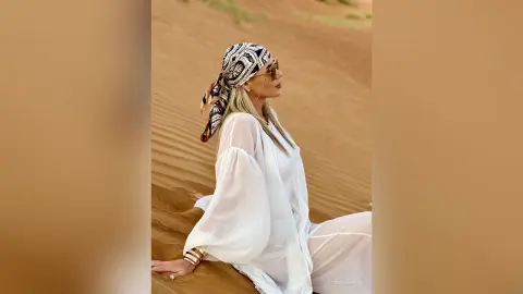 TheaSiena — Stylish woman in white bohemian outfit and patterned headscarf relaxing in a sunny desert sand dune setting.