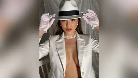 Elegant woman in a white blazer and pearl necklace wearing a white fedora and gloves in a bright studio setting.