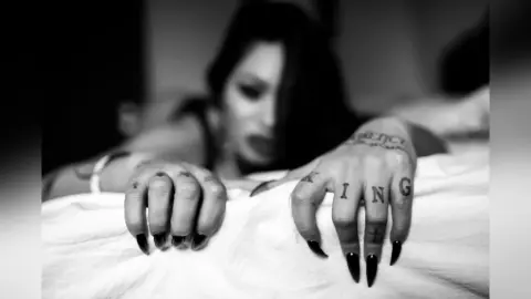 DeeaAnderson rakes her tattooed hands across white bedsheets, looking toward the camera with a heavy-lidded gaze.