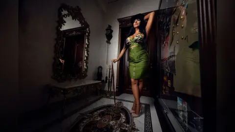 MissyJolie stands in a dimly lit room wearing a tight green lace bra and skirt, posing next to an ornate gold mirror.