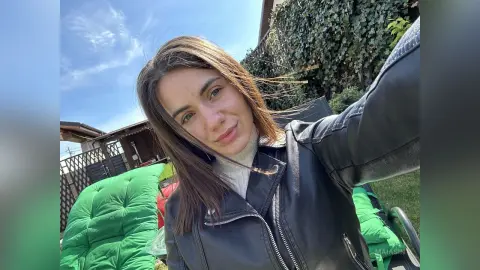 Smiling young woman in a black leather jacket relaxing outdoors on a green lounge chair under a blue sky.