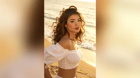 Beautiful woman in a white off-the-shoulder blouse posing on a sunny beach at sunset with a serene mood.