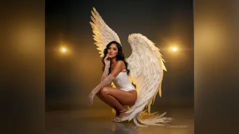 Elegant woman in white corset and lace gloves posing with large angel wings in a moody studio setting.