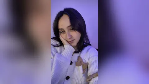 Smiling young woman in a cozy white knit cardigan posing in soft purple ambient lighting.
