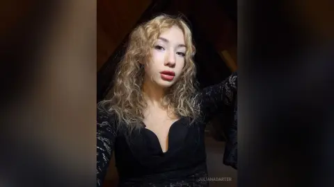 Blonde woman in a black lace long-sleeve top posing with a moody aesthetic in a dimly lit room.